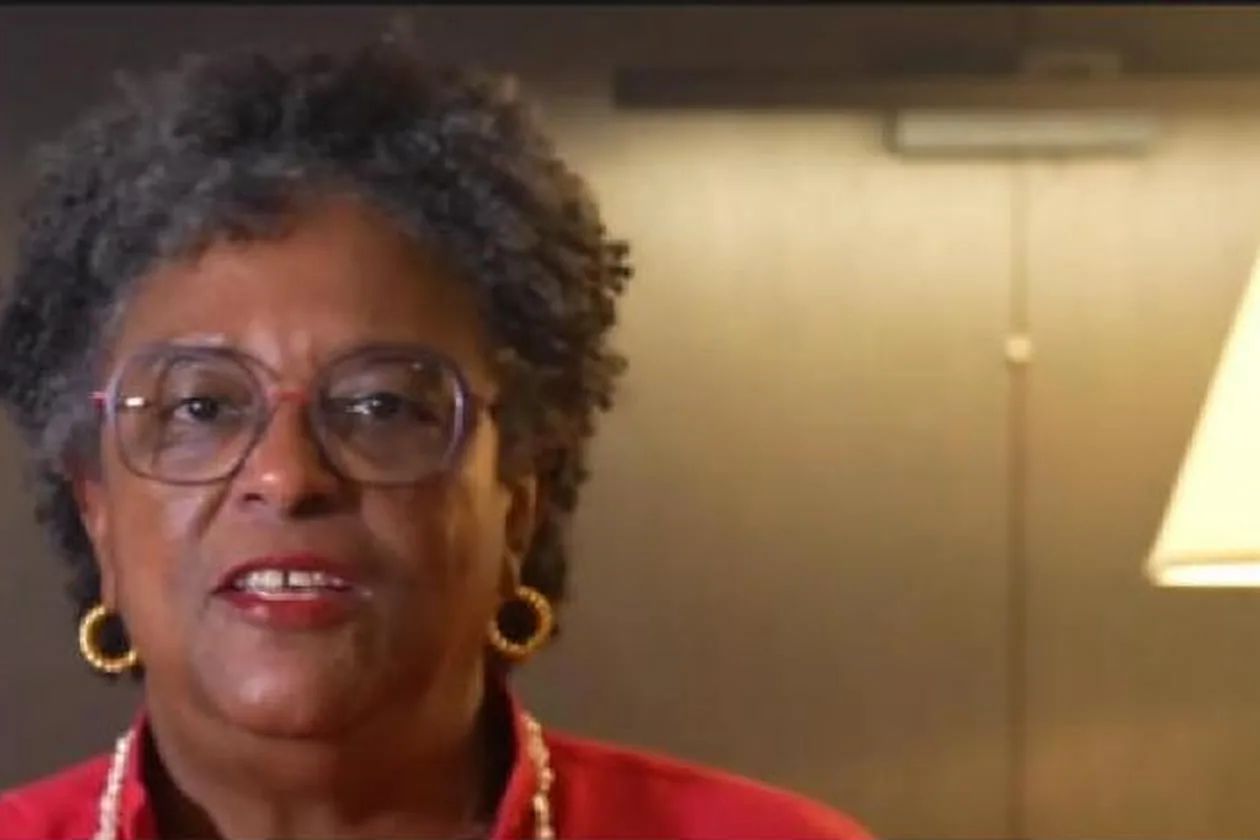 Mia Mottley, wearing glasses, a red top and a pearl necklace, speaking to camera in front of a lamp.