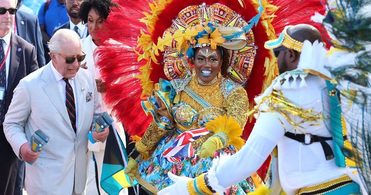 King Charles in sunglasses smiles as a Junkanoo dancer in a red and gold feathered headdress and floral costume, holding a small Union flag, dances in front of him