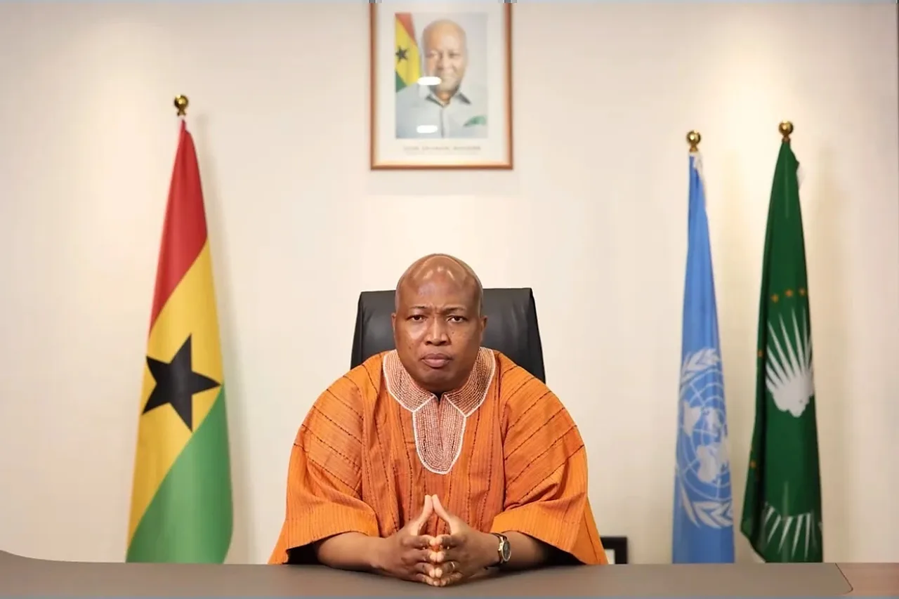 Samuel Okudzeto Ablakwa in an orange smock seated at a desk with his hands clasped, between the flags of Ghana, the United Nations and the African Union.